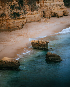 View of golden cliffs meet the turquoise sea, waves gently kiss the sandy shore, and a solitary figure graces the beach, Portimao, Faro, Portugal.