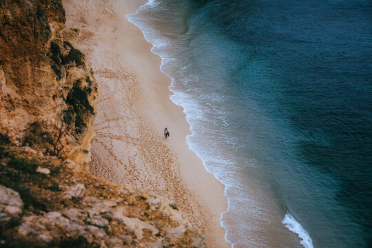 View of golden sands meet the turquoise embrace of the ocean, as cliffs rise majestically, dwarfing tiny figures strolling along the shore, Portimao, Faro, Portugal.