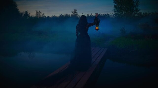 gothic woman hand hold lantern lamp magic warm light, lady walking across bridge over river lake dark fog mist night sky nature way. long black retro style black dress. Fantasy scared girl looks back.