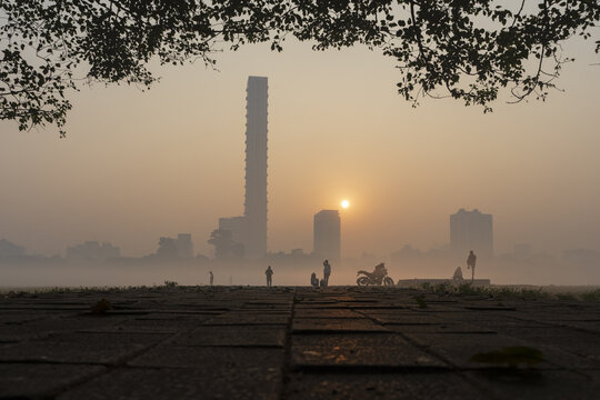 View of a hazy sunrise casting a warm glow over the silhouetted cityscape and the people enjoying the peaceful morning, Kolkata, West Bengal, India.