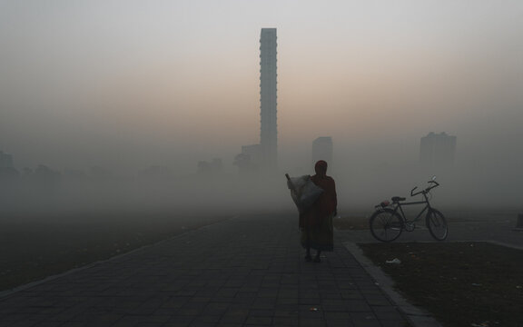 View of a solitary figure walks along a stone path, shrouded in a dense fog, with a bicycle nearby and a towering skyscraper in the distance, Kolkata, West Bengal, India.