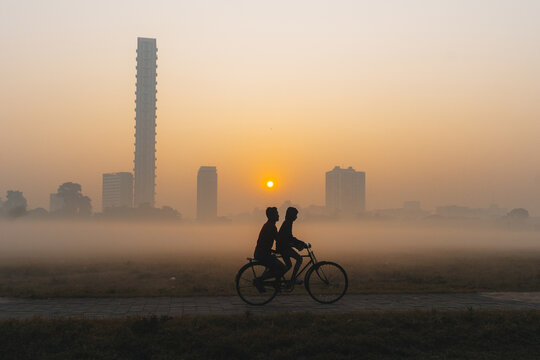 View of silhouettes of two people riding a bicycle through a misty landscape, framed by modern skyscrapers under a glowing sunrise, Kolkata, West Bengal, India.
