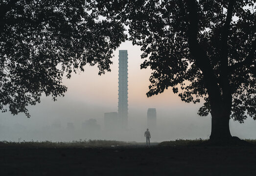 View of a solitary figure stands amidst the ethereal morning mist, framed by the dark silhouettes of trees, with the city's tall buildings fading into the horizon, Kolkata, West Bengal, India.