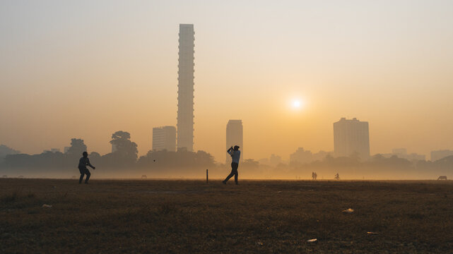 View of cricket players silhouetted in the early morning light against the backdrop of Kolkata's skyline, shrouded in a golden haze, Kolkata, West Bengal, India.