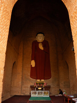 View of a serene Buddha statue stands tall in a temple, the red robes contrasting with the aged brickwork, Old Bagan, Mandalay Region, Myanmar Burma.