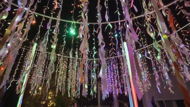 Numerous colorful ribbons and string lights hanging from a circular frame, creating a festive and illuminated canopy at night.