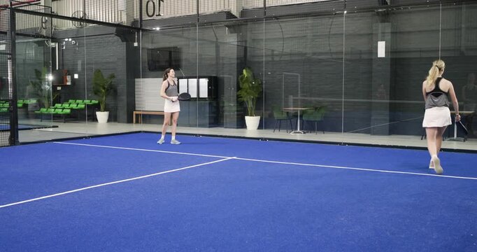 After ball crossing net, two female athletes volleying then rallying with rackets on padel court