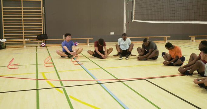 Diverse male players and coach placing cones and initiating seated hamstring stretches for warmup