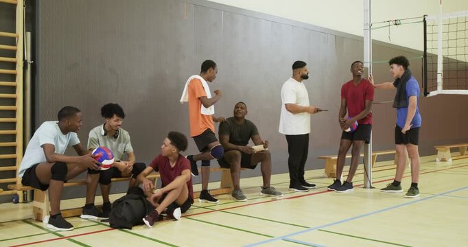Diverse male team sitting on bench in school gym during timeout, passing volleyball, checking phone