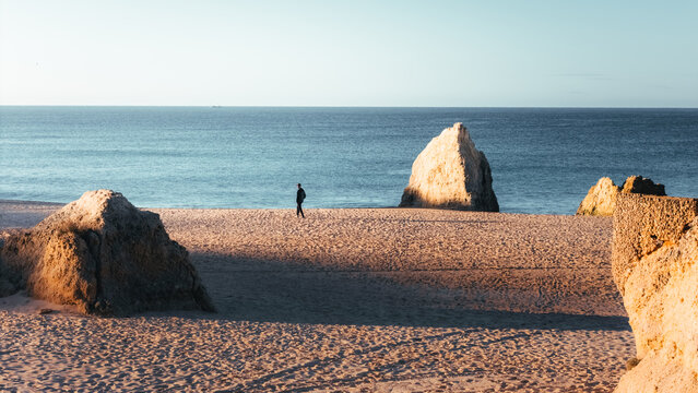 Aerial view of a solitary figure strolls across the sun-kissed sands of Praia dos Tres Irmaos, framed by monolithic rock formations against the tranquil sea, Alvor, Faro, Portugal.