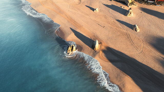 Aerial view of golden sands meeting the turquoise sea, punctuated by the dramatic rock formations of Praia dos Tres Irmaos, Alvor, Faro, Portugal.