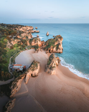Aerial view of the golden sands of Praia dos Tres Irmaos embrace jagged cliffs as turquoise waves crash gently against the shore, Alvor, Faro, Portugal.
