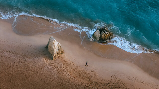 Aerial view of the golden sands meeting the turquoise sea at Praia dos Tres Irmaos, where rugged rock formations stand against the rhythmic waves, Alvor, Faro, Portugal.