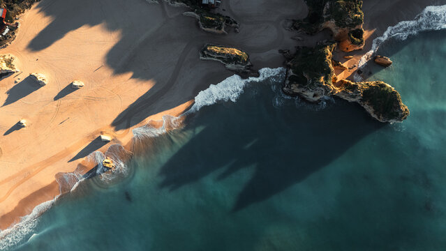 Aerial view of golden sands meeting the turquoise sea at Praia dos Tres Irmaos, cliffs casting long shadows onto the beach, Alvor, Faro, Portugal.