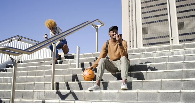 Diverse pair on urban stairs, man with basketball speaking during call, woman sitting with coffee