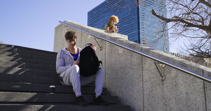 Man, woman on stairs, he checking watch pulling paperbag from backpack eating while she using phone