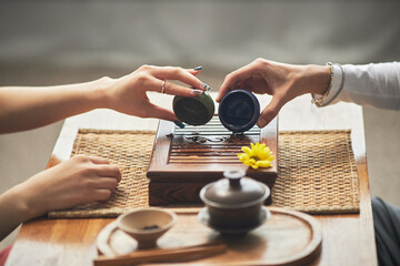 Asian young adult woman and Asian young adult man performing traditional tea ceremony, both hands holding teacups over wooden tea tray, engaging in cultural ritual together