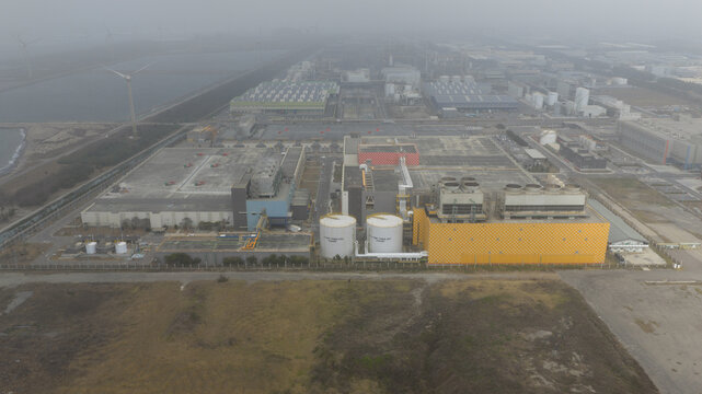 Aerial view of industrial buildings casting long shadows under a hazy sky, with wind turbines faintly visible in the distance, Changhua County, Taiwan.