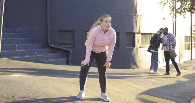Woman in pink hoodie jogging, stopping, catching breath at curved wall, checking watch, tying shoe