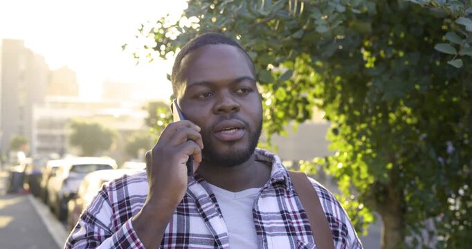 Receiving call, African American man holding phone to right ear, smiling on sidewalk with bag strap
