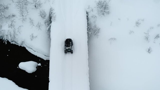 Aerial top down overview of offroad tank track vehicle crossing narrow snow covered bridge over river