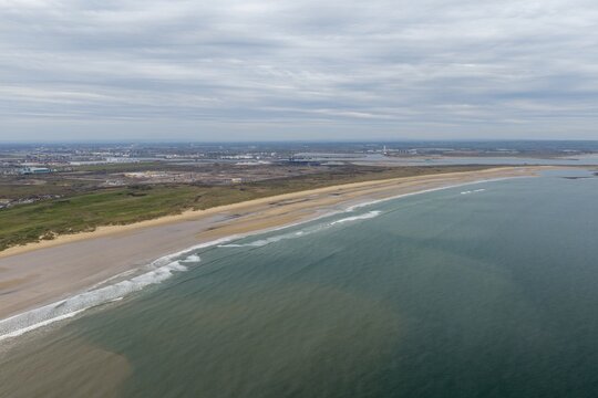 Aerial coastline near Redcar Middlesbrough showing beach sea waves and distant industrial area with port buildings across a wide landscape scene along the shore in the spring time