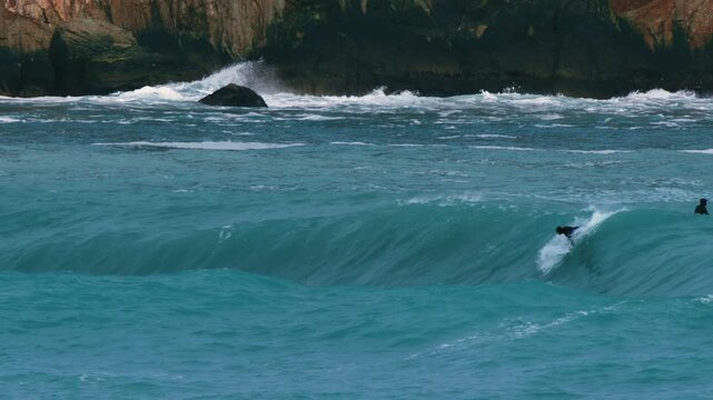 Surfer dropping into powerful ocean wave near rocky coastline, slow motion paddle in and bail off wave