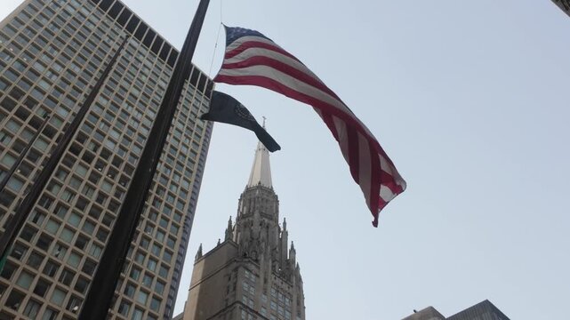 A large American Flag and a POW MIA flag blowing in the wind in front of skyscrapers in the city of Chicago
