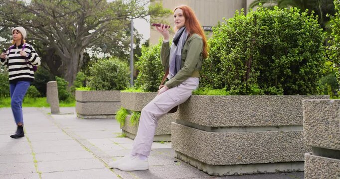 Mid-adult woman holding smartphone to mouth, talking on call while sitting on concrete planter