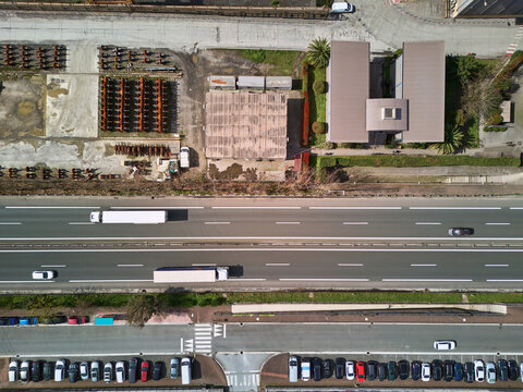 Olaberria, Spain - 09 March 2026: Aerial view of a highway bisecting the industrial landscape, with trucks traversing the grey asphalt and cars parked in neat rows below, all under a muted sky.