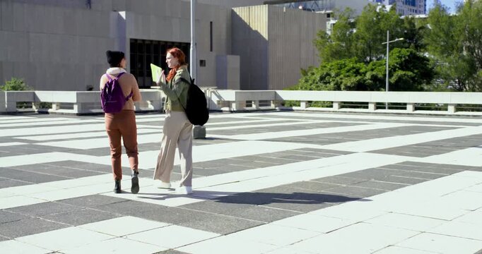 Female friends walking plaza, red-haired woman extending yellow notes to companion with purple pack