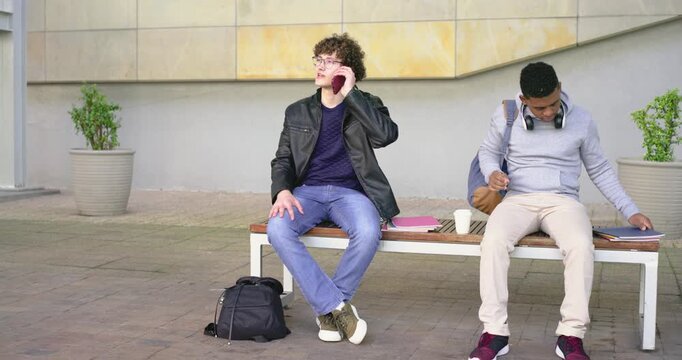 Diverse male students meeting on bench, one talking on phone, other arriving with cup shaking hands