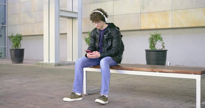 Curly-haired man lifting white headphones tapping red smartphone on plaza bench for music