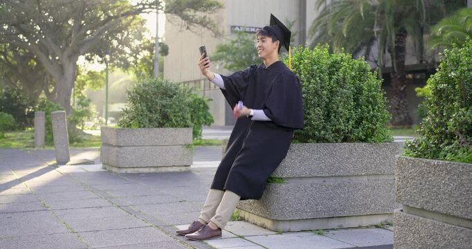 Postgrad Asian man wearing cap gown plaza holding phone and degree taking selfie pausing for walker