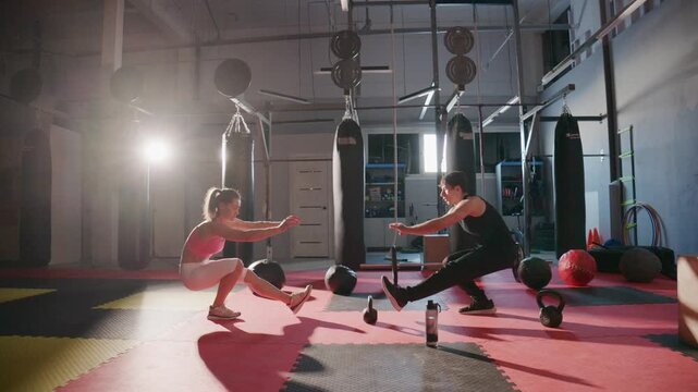 Young couple trains together in dim gym. Sunlight streams through window, casting bold shadows. They move with focused energy, pushing limits side by side