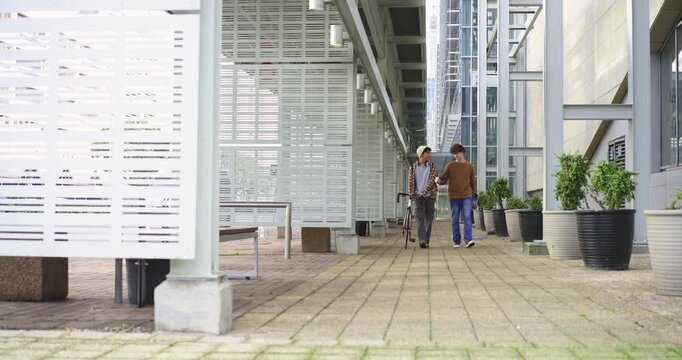 Asian man and male friend entering walkway, walking toward camera pushing bike and discussing notes