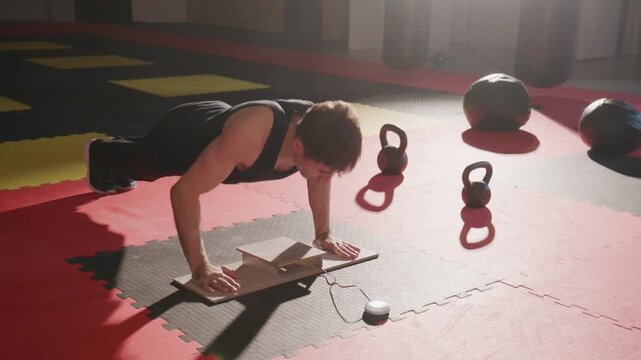 Young man pushes hard on a wooden board. Sunlight streams through a gym's windows. Kettles, balls, and mats surround him. He strains, focused, determined to push through. Perfect for fitness, strength