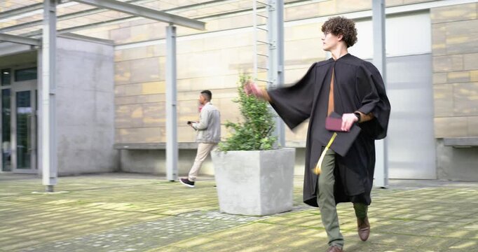 Adult male graduate in gown walking campus holding cap and folder waving to passerby entering doors
