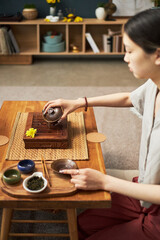 Young adult Asian woman performing traditional tea ceremony, pouring tea from teapot into cup on wooden table, sitting indoors with tea utensils and loose leaf tea visible