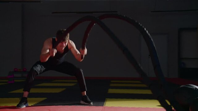 Athlete grips heavy ropes with intense focus. His stance shows power and determination. He pulls with force, muscles straining. Dark gym lights highlight his effort