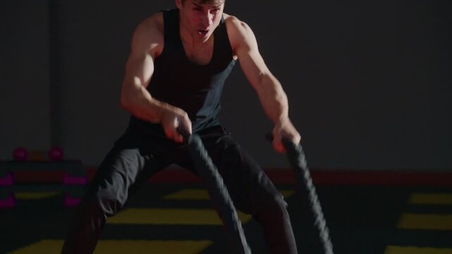 Young man strains, pushing hard in dark room. His face shows intense effort, sweat glistening on skin. He crouches low, focused, ready to explode with power. Ideal for fitness, strength