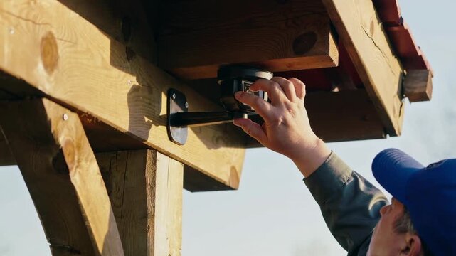 Outdoor security camera install white technician mounting bracket under wooden pergola, hands adjusting sensor and lens with drill and screws, daytime residential porch, contractor uniform and cap