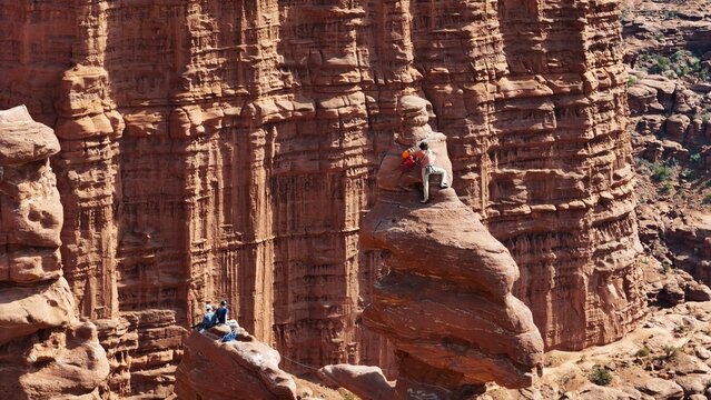 Aerial view of climbers daringly perched atop a towering sandstone spire, its rust-colored rock contrasting against the vast canyon, Moab, Utah, United States.