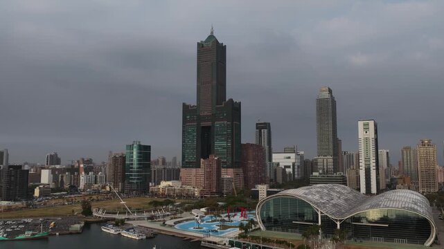 Overcast aerial view of the modern kaohsiung city skyline, featuring the iconic 85 sky tower, the music center, and luxury yachts docked in the bustling harbor of love river bay in taiwan