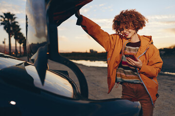 Fototapeta premium Smiling woman with curly hair wearing a rainbow sweater and orange jacket stands by a car at sunset holding a smartphone, enjoying a relaxed lifestyle moment outdoors.