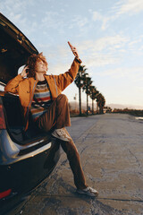 Fototapeta premium Young woman with curly hair wearing rainbow sweater and brown jacket sits in car trunk smiling and taking selfie with smartphone on sunny day near palm trees on empty road.