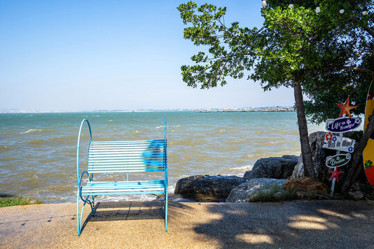 Blue metal bench facing a windy sea under a clear sky, with a tree, decorative signs and surfboard on the side, creating a relaxing seaside atmosphere.