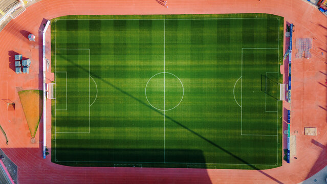 Novi Sad, Serbia - 11 March 2026: Aerial view of a vibrant green football field contrasts sharply with the surrounding red running track, bathed in sunlight.