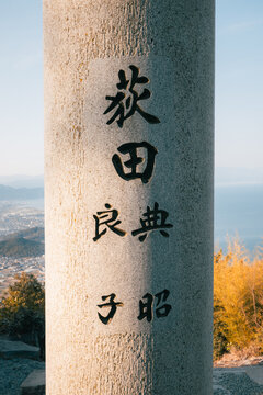 View of weathered stone pillar etched with Japanese calligraphy, standing tall against a backdrop of the shimmering Seto Inland Sea, Kanonji, Kagawa, Japan.