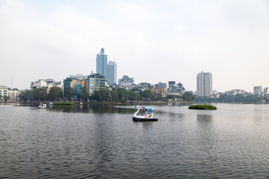 View of swan paddle boats drift on the tranquil lake under a soft sky, with city buildings and trees lining the distant shore, Hanoi, Ha Noi, Vietnam.
